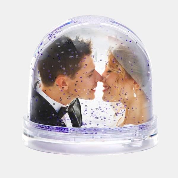 Die Schneekugel mit violettem Glitzer zeigt ein Brautpaar kurz vor einem Kuss, das sich liebevoll ansieht.