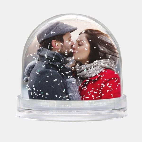 Die Schneekugel mit Schnee zeigt ein Paar im Wintermantel, das sich liebevoll vor einem Kuss anschaut.